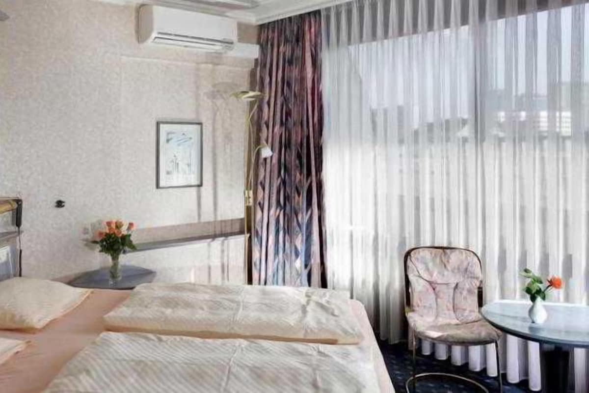 A hotel room with a bed, a chair, a small table, and a window with curtains.