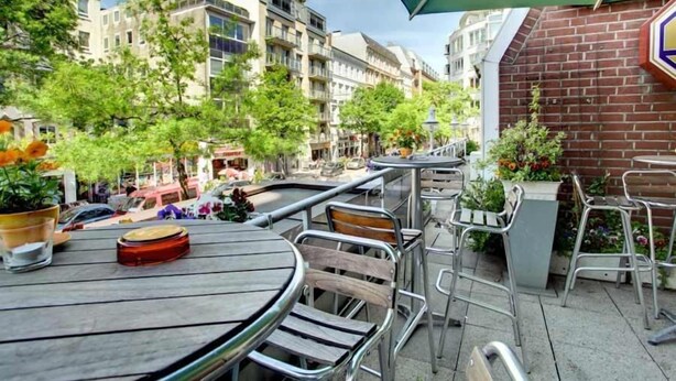 Un espace extérieur aménagé avec des tables rondes et des chaises en métal, entouré d’un mur de briques et de verdure.