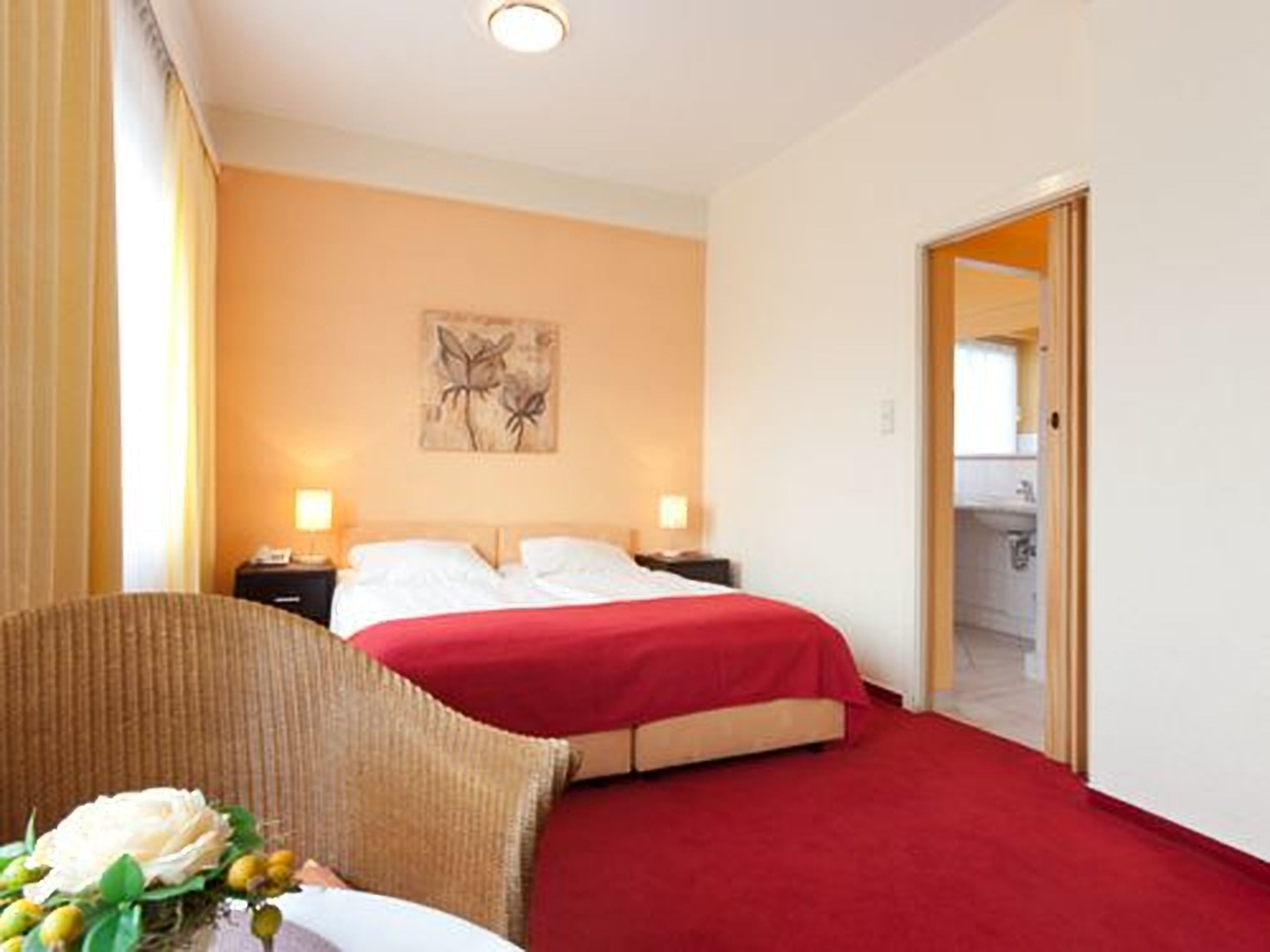 A hotel room with a bed, two bedside tables, a chair, a red carpet, and a painting on the wall.