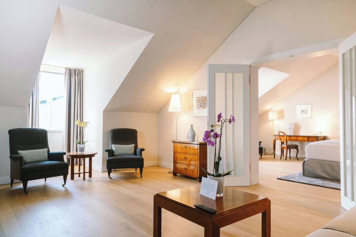 A modern hotel room with a high ceiling, wooden flooring, a sofa, two armchairs, a small table, a desk, a chair, a dresser, and a bed.