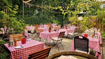 Un patio al aire libre con mesas cubiertas con manteles a cuadros rojos y blancos, sillas de madera y luces colgantes.