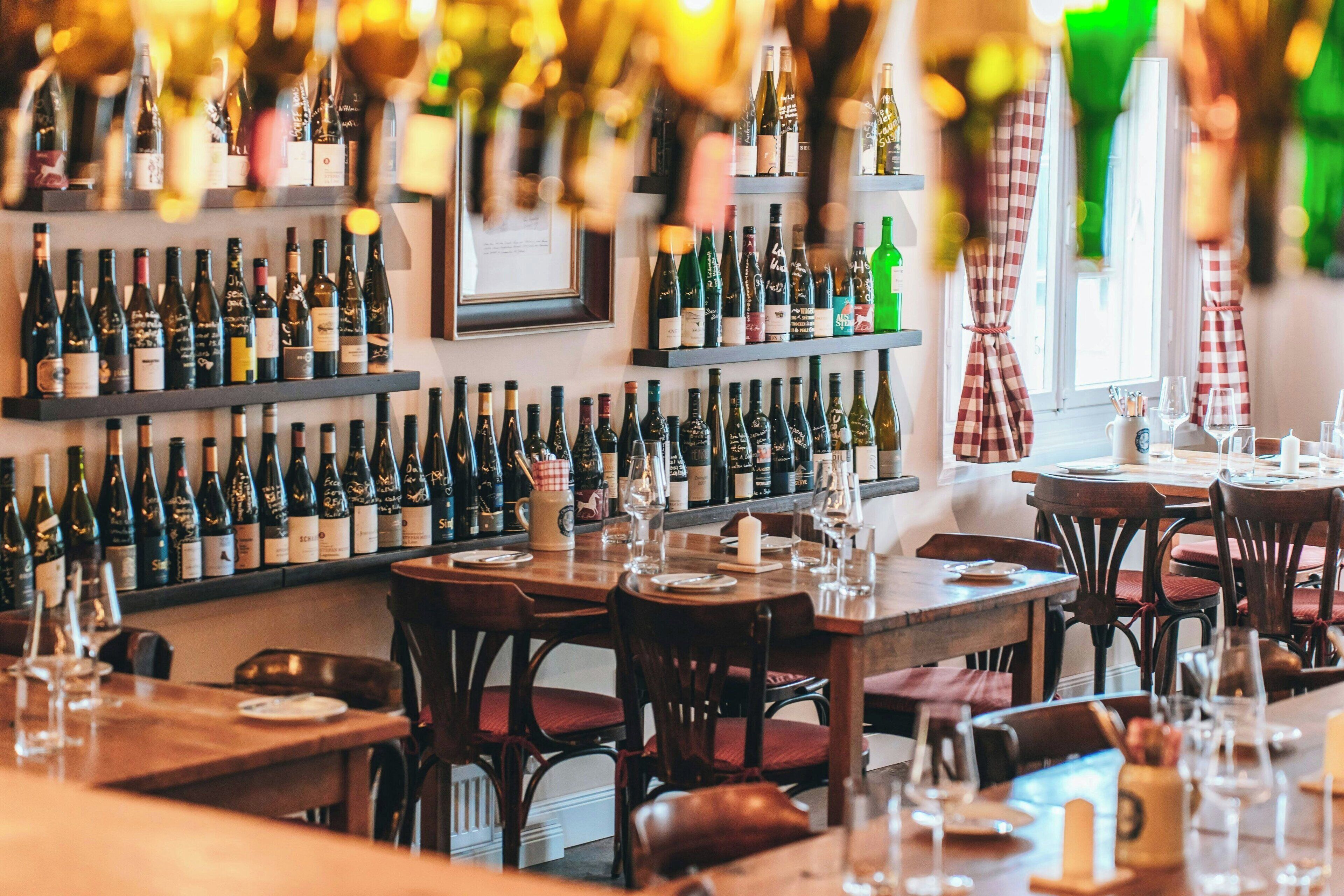 A restaurant with multiple tables and chairs, shelves with wine bottles, and a window with curtains.