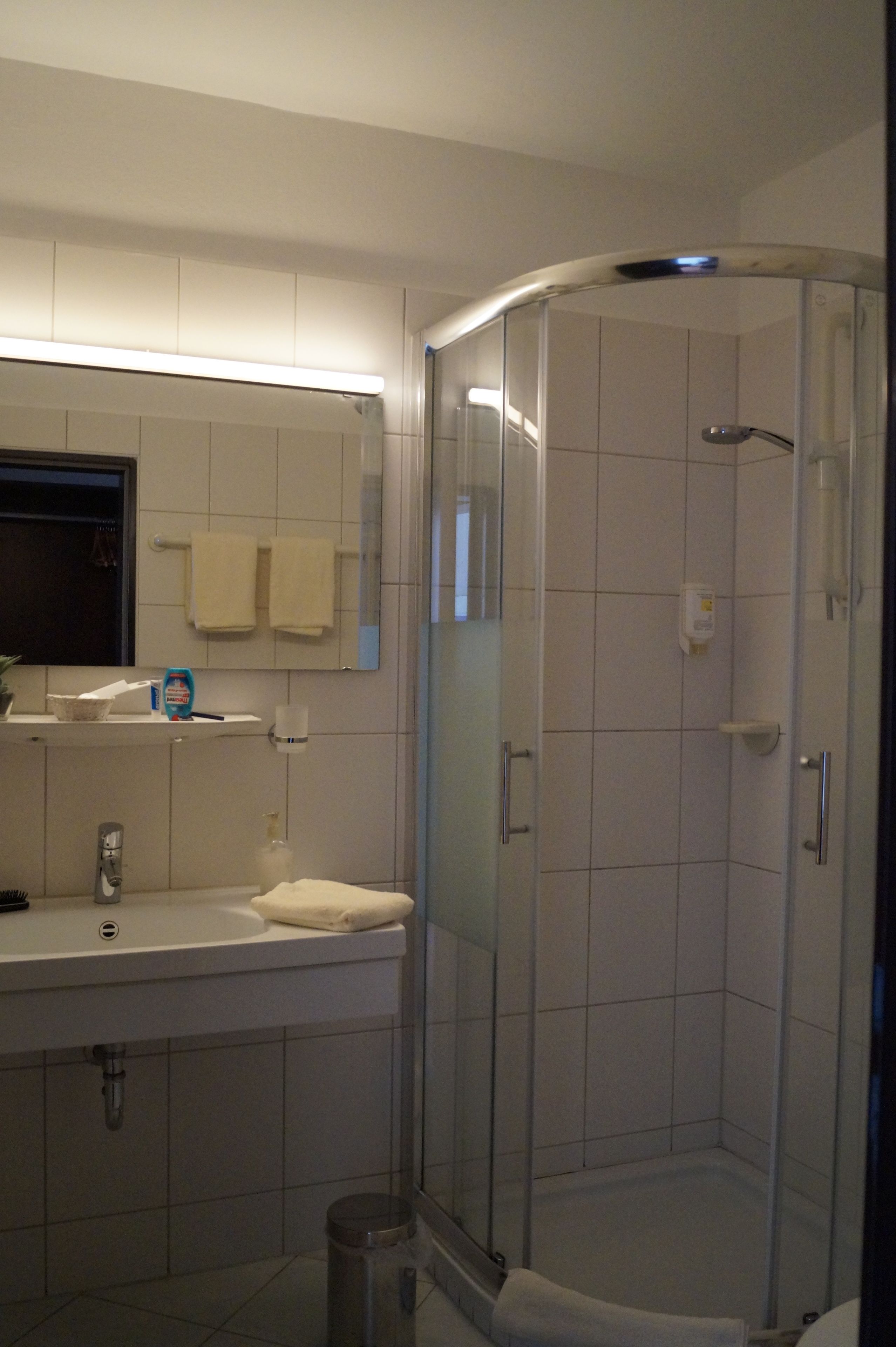 A bathroom with a glass shower enclosure, white tiles, a towel rack, and a window.