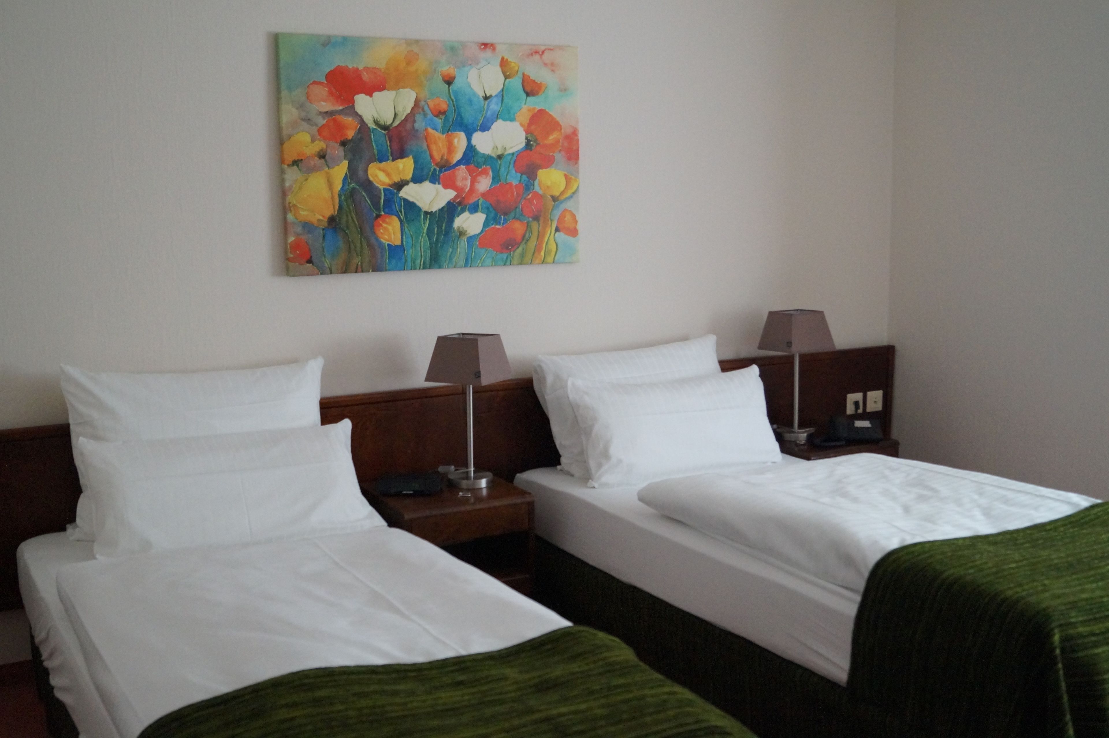 A hotel room with two single beds, a nightstand with a lamp, and a painting of flowers on the wall.