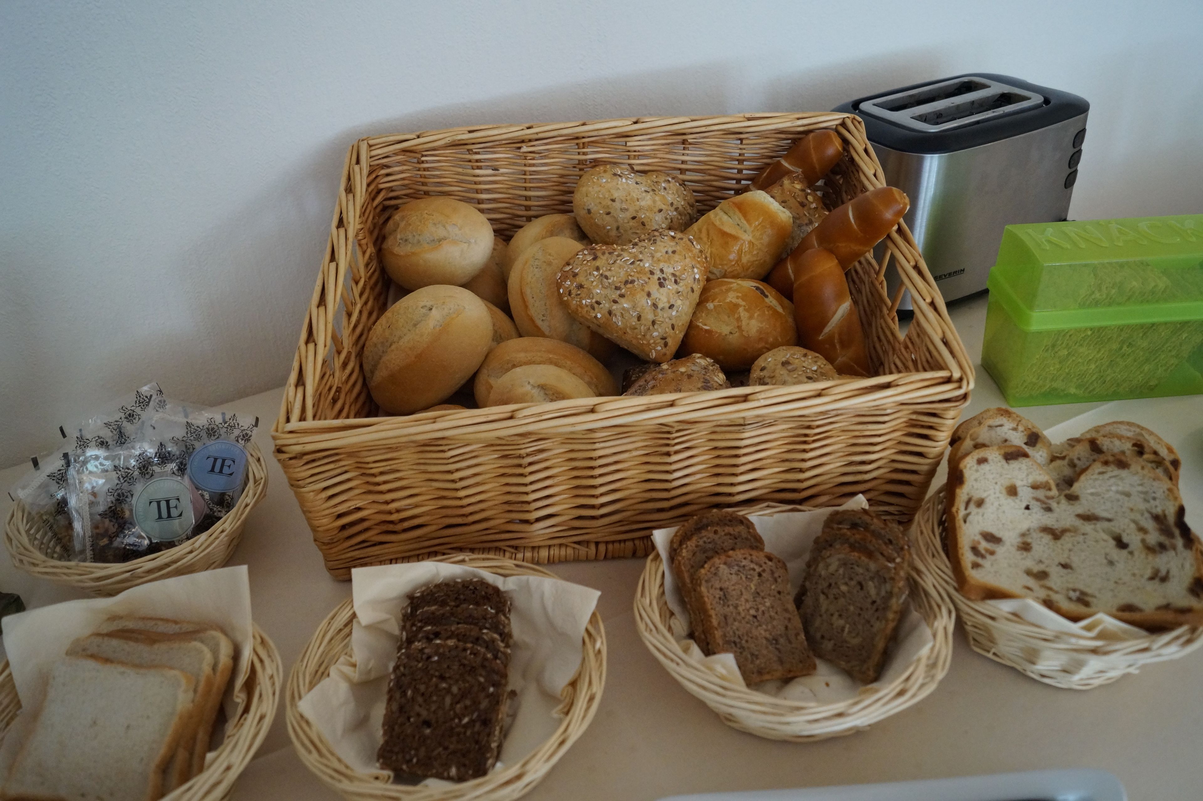 Variedad de panes y pasteles en cestas de mimbre, un tostador y una caja verde sobre una mesa.