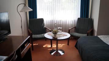 Una habitació d'hotel amb un llit, una taula de treball, una cadira, una taula petita amb un ram de flors i una làmpada.