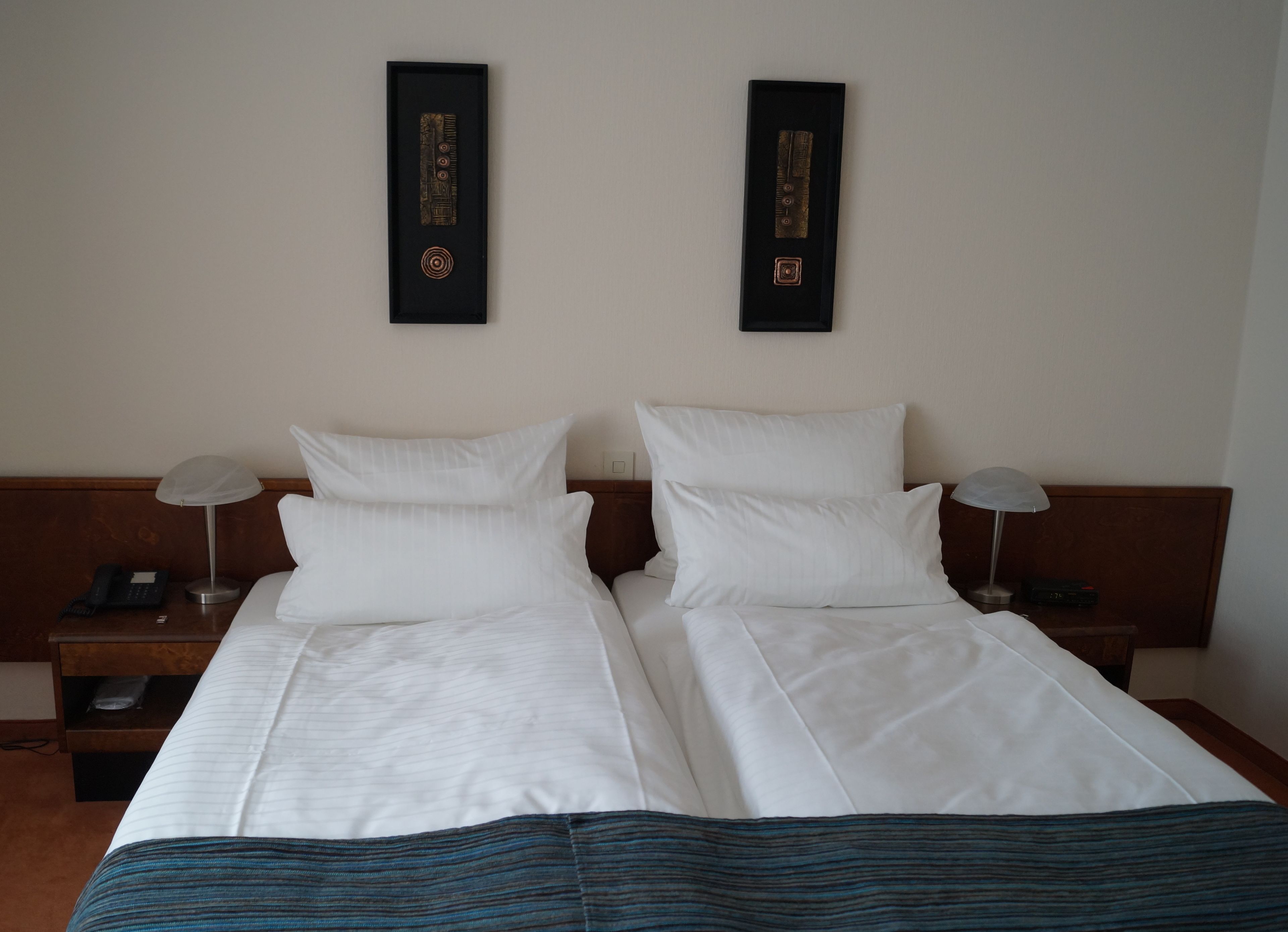 Una cama doble con sábanas blancas, dos mesitas de noche con lámparas y dos adornos en la pared.