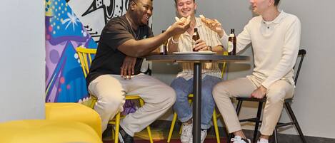 Three people seated at a table with pizza, bottles, and a colorful wall mural in the background.