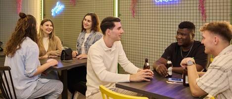 A group of people sitting around a table in a room with grid-patterned walls, neon signs, and hanging plants.