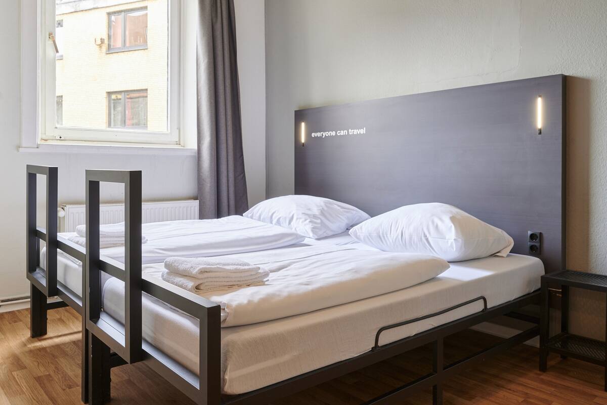 A double bed with white linens, a grey headboard with a light strip, and a window with curtains.