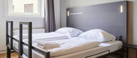Cama doble con ropa blanca, cabecera gris con iluminación y ventana con cortinas.