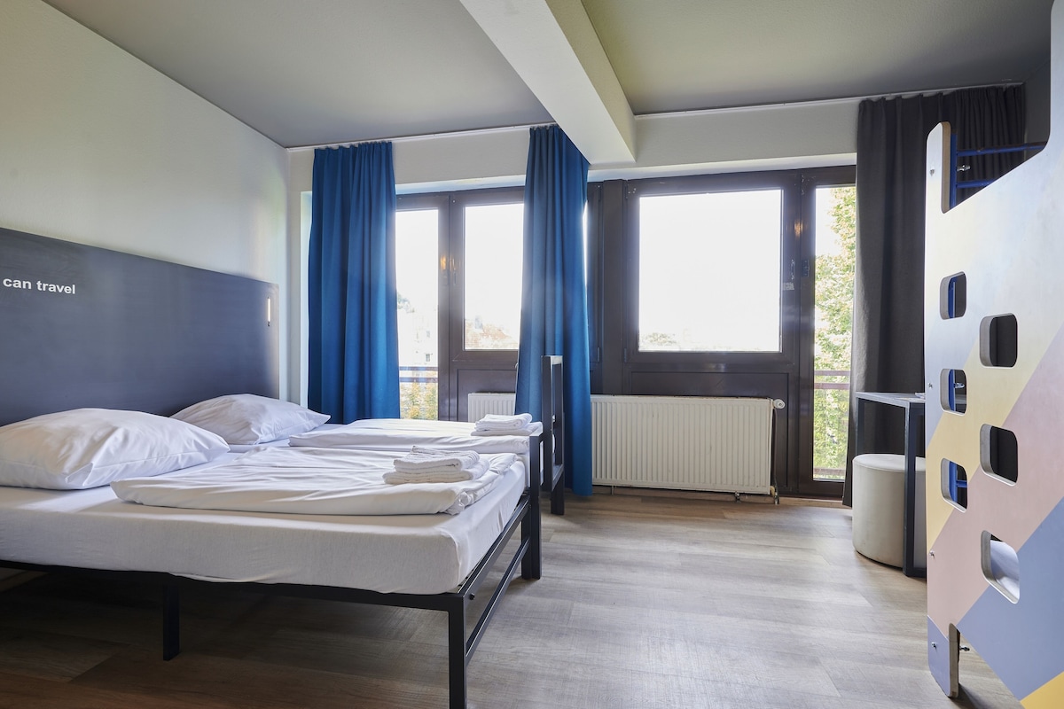 Quarto de hotel com cama, janela com cortinas, radiador e unidade de armazenamento na parede.