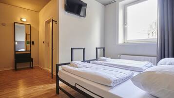Ein Hotelzimmer mit zwei Betten, einem an der Wand befestigten Fernseher, einem Fenster mit Vorhängen und einer Bank in der Ecke.