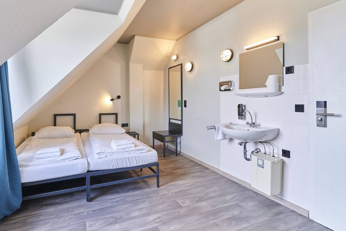 A modern hotel room with a sloped ceiling, two beds with neatly made white linens, a large mirror, a wall-mounted sink, and a small table.