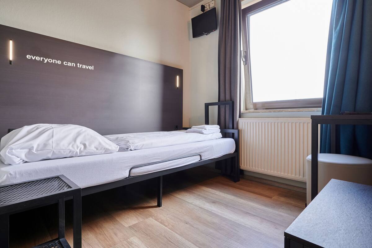 A small, modern hotel room with a single bed, a desk, and a window with curtains.