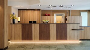 A modern reception desk with a wooden finish, a bowl of fruit, and a vase of flowers. - Leonardo Hotel & Residenz München (Munich)