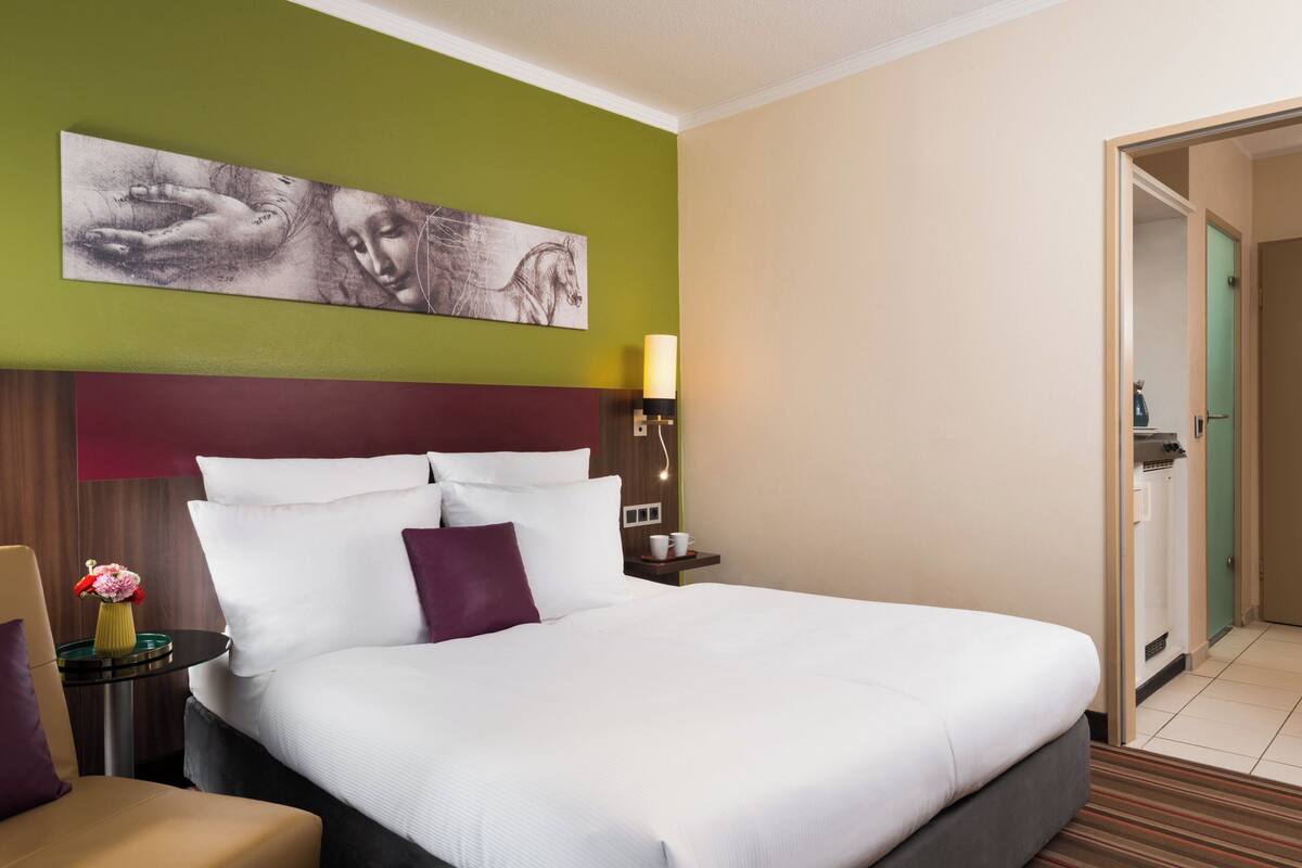 A hotel room with a large bed, a headboard, a nightstand with a lamp, a small table with a vase of flowers, and a painting on the wall.