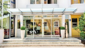 A hotel entrance with a glass door, a covered waiting area, and a yellow door. - Leonardo Hotel & Residenz München (Munich)