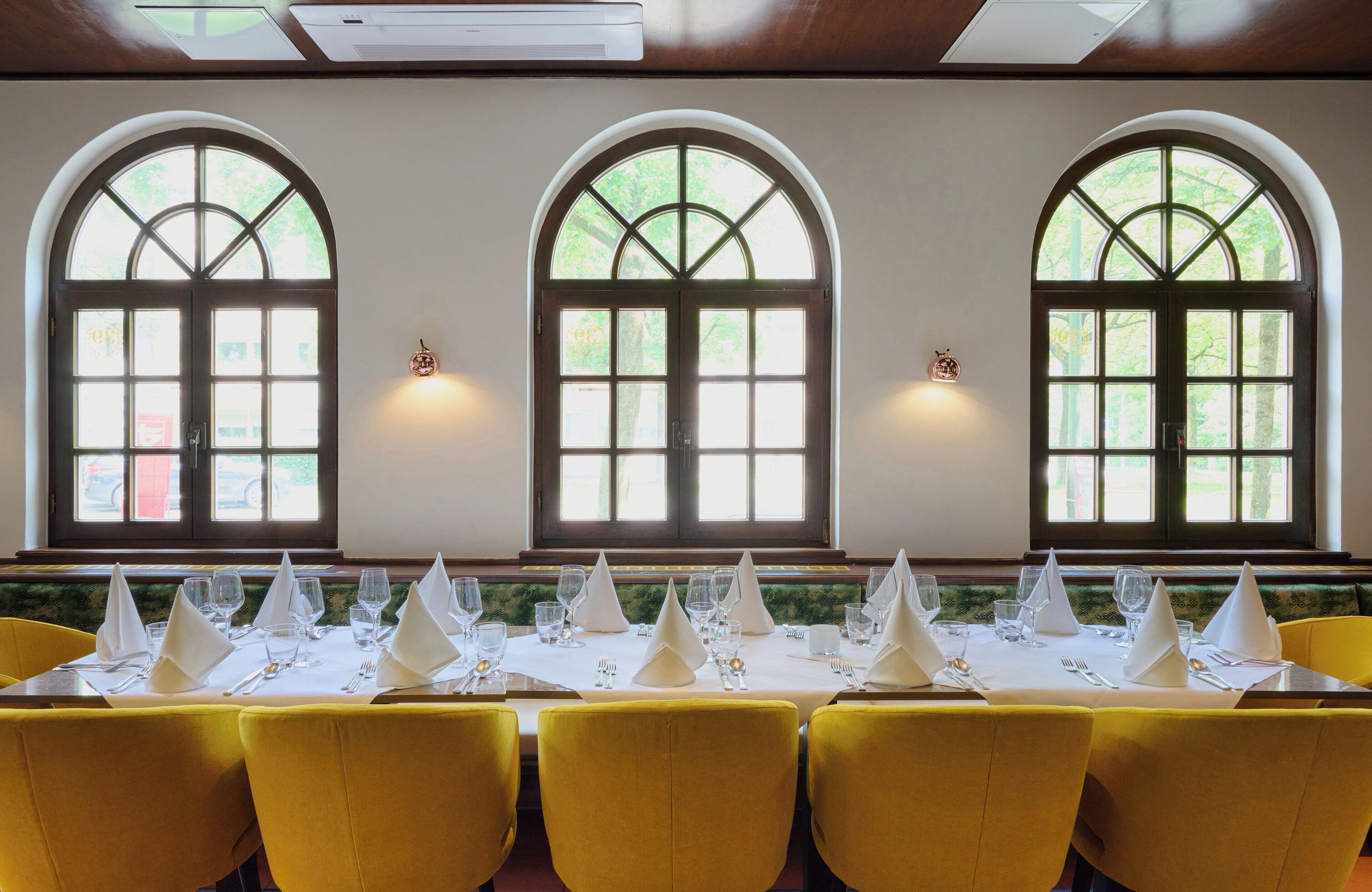 A dining area with yellow chairs and tables, large arched windows, and a white tablecloth.