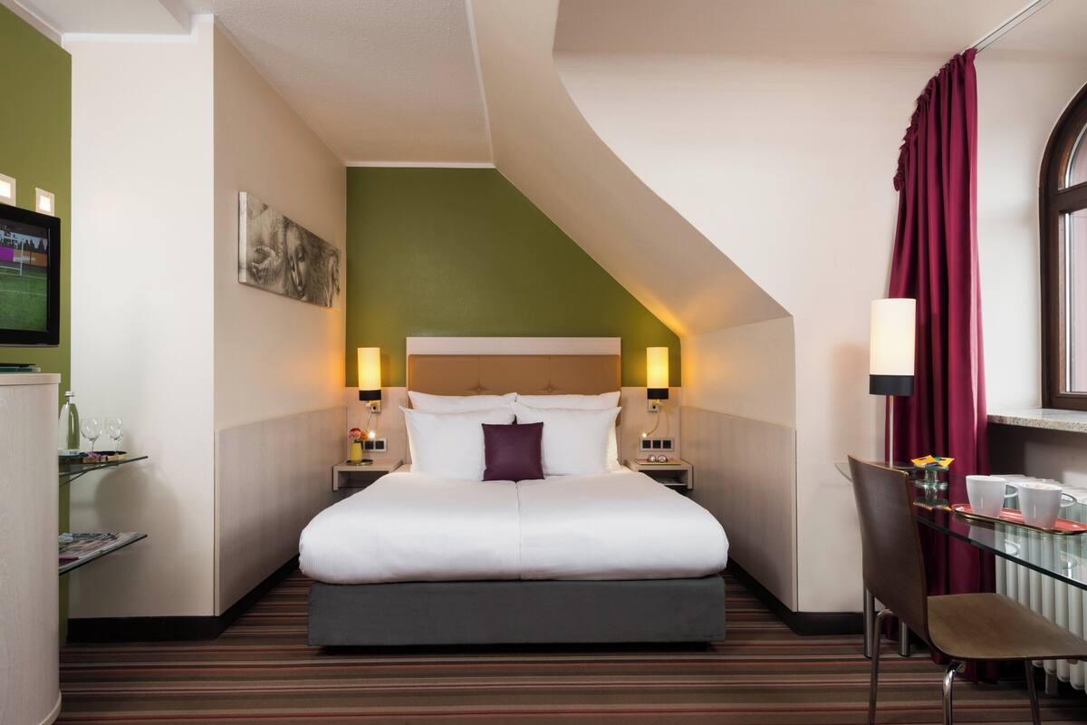 Una habitación de hotel con una cama grande, un escritorio con silla, televisión y ventana con cortinas.