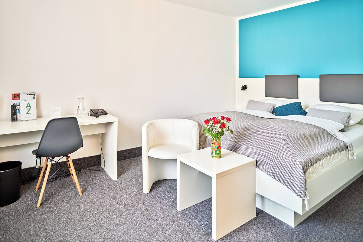 A hotel room with a bed, a desk, a chair, a small table, and a vase with flowers.