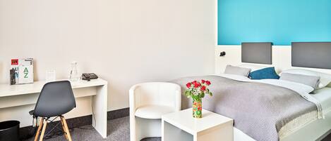 A hotel room with a bed, a desk, a chair, a small table, and a vase with flowers.