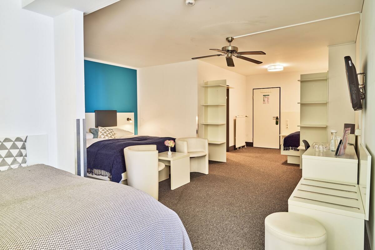 A hotel room with a bed, a desk, a chair, a ceiling fan, and a door.