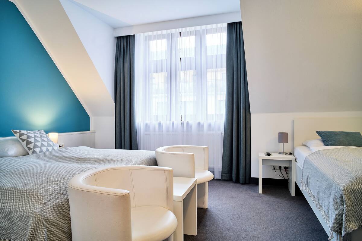 A hotel room with a bed, two chairs, a nightstand, and a window with curtains.
