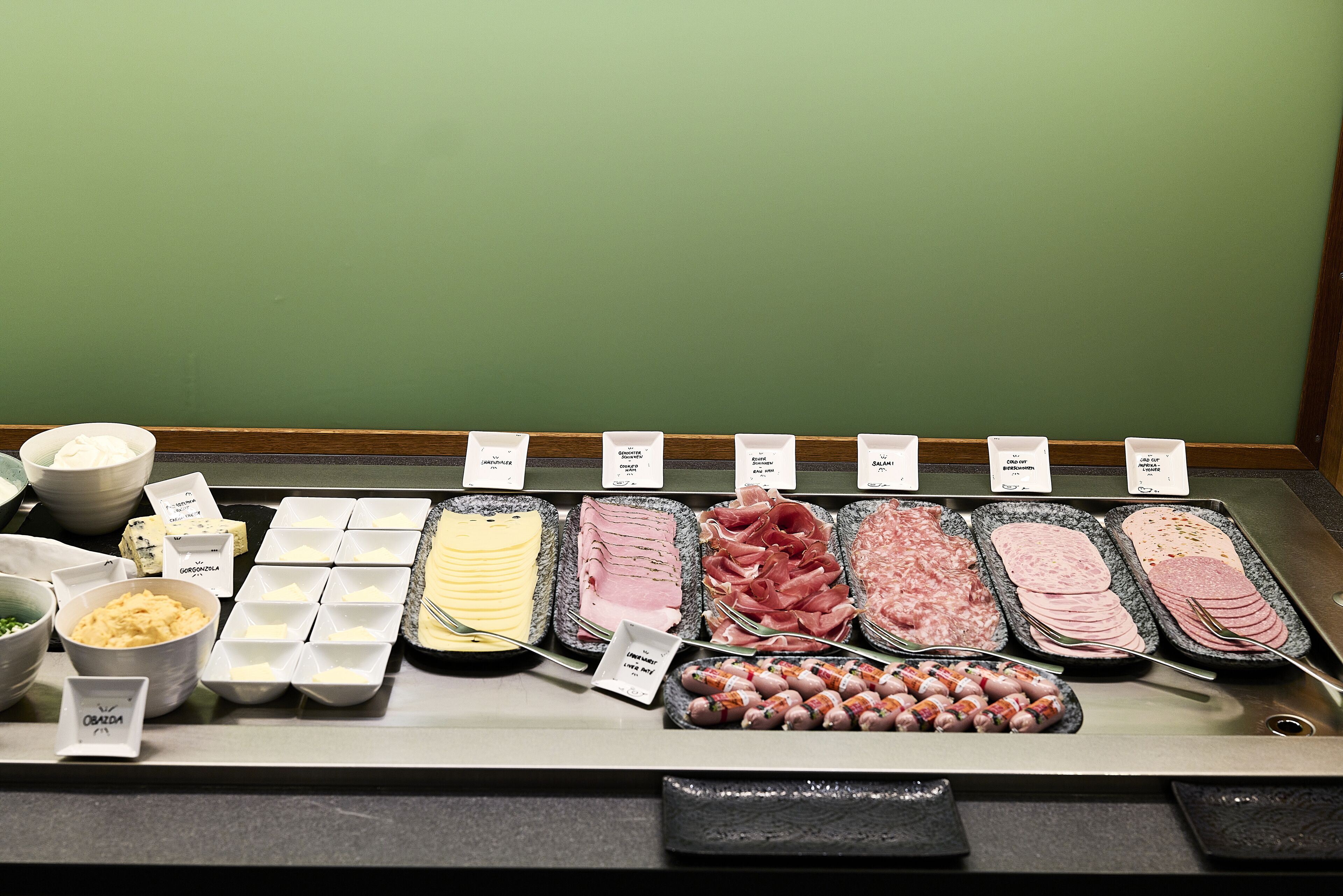A deli counter with various types of deli meats and cheeses displayed in trays, each labeled with its name.