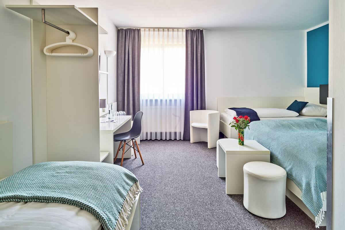 A hotel room with a bed, a desk, a chair, a small table, and a hanging coat rack.