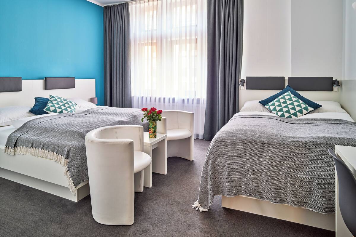 A modern hotel room with two beds, a small table with a vase of flowers, a chair, and a view of the city through large windows.