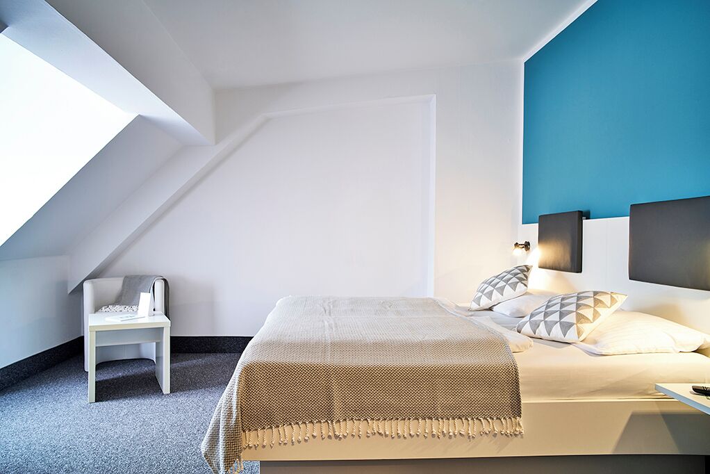 A modern bedroom with a sloped ceiling, a bed with a beige blanket, a small white chair, and a wall-mounted TV.