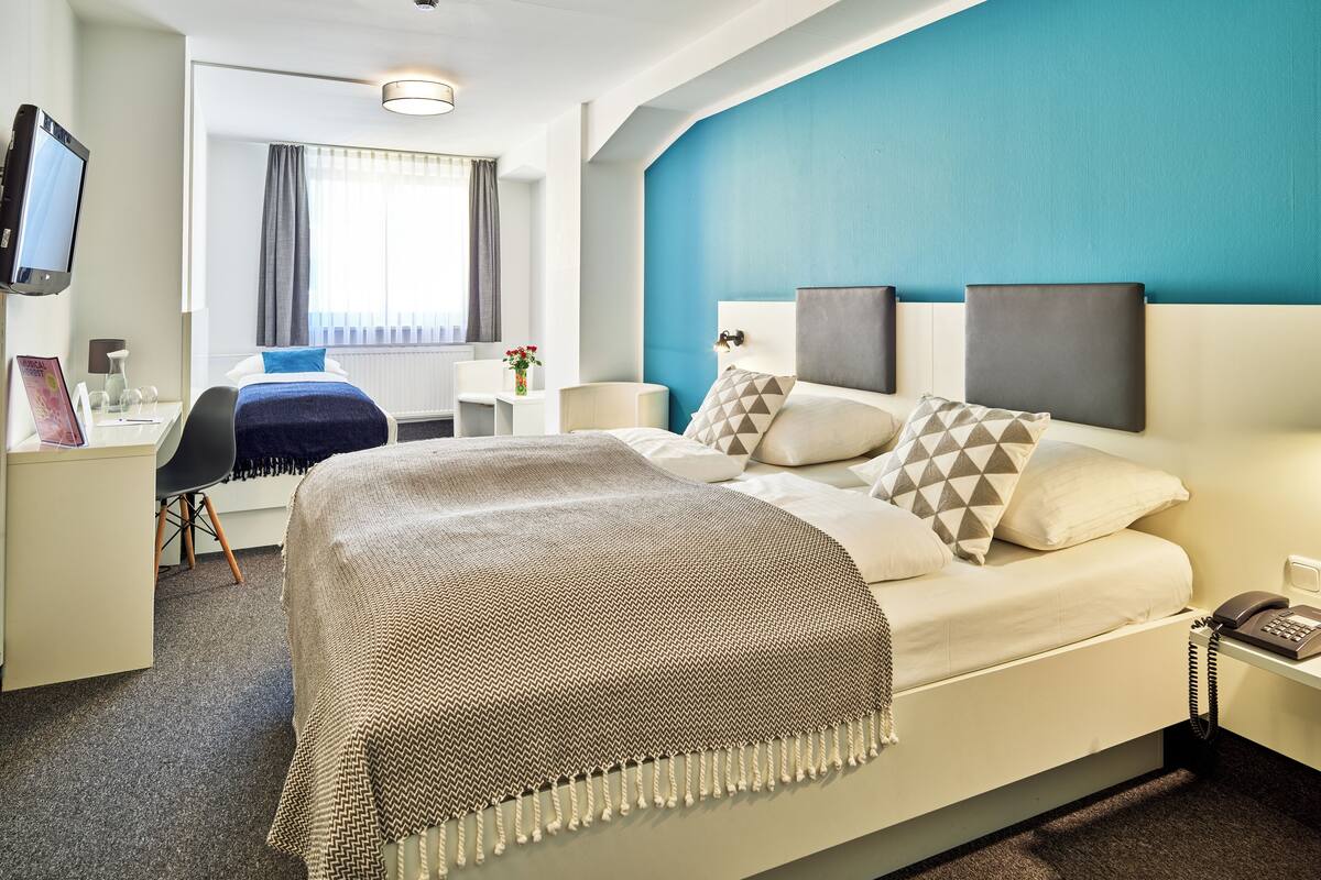 A hotel room with a bed, bedside tables, a desk, a chair, a TV, and a window with curtains.