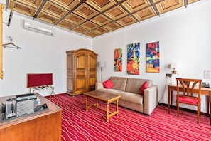 A hotel room with a red carpet, a sofa, a wooden cabinet, a desk with a chair, and a ceiling with a decorative pattern.