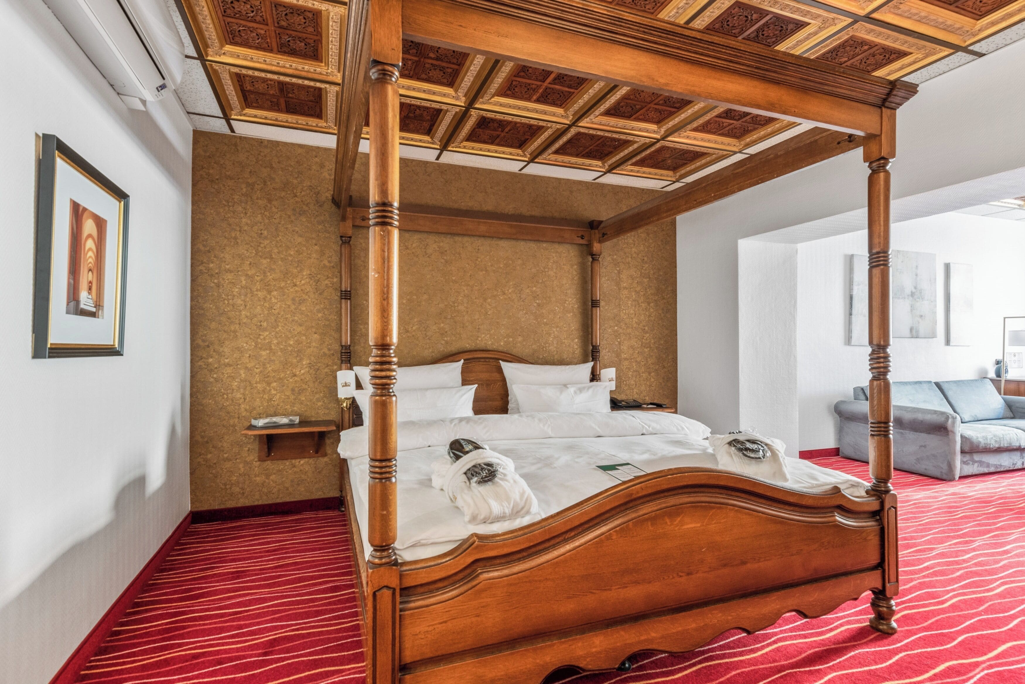 Una cama con dosel de estructura de madera y ropa de cama blanca, suelo cubierto de alfombra roja y techo con diseño decorativo.