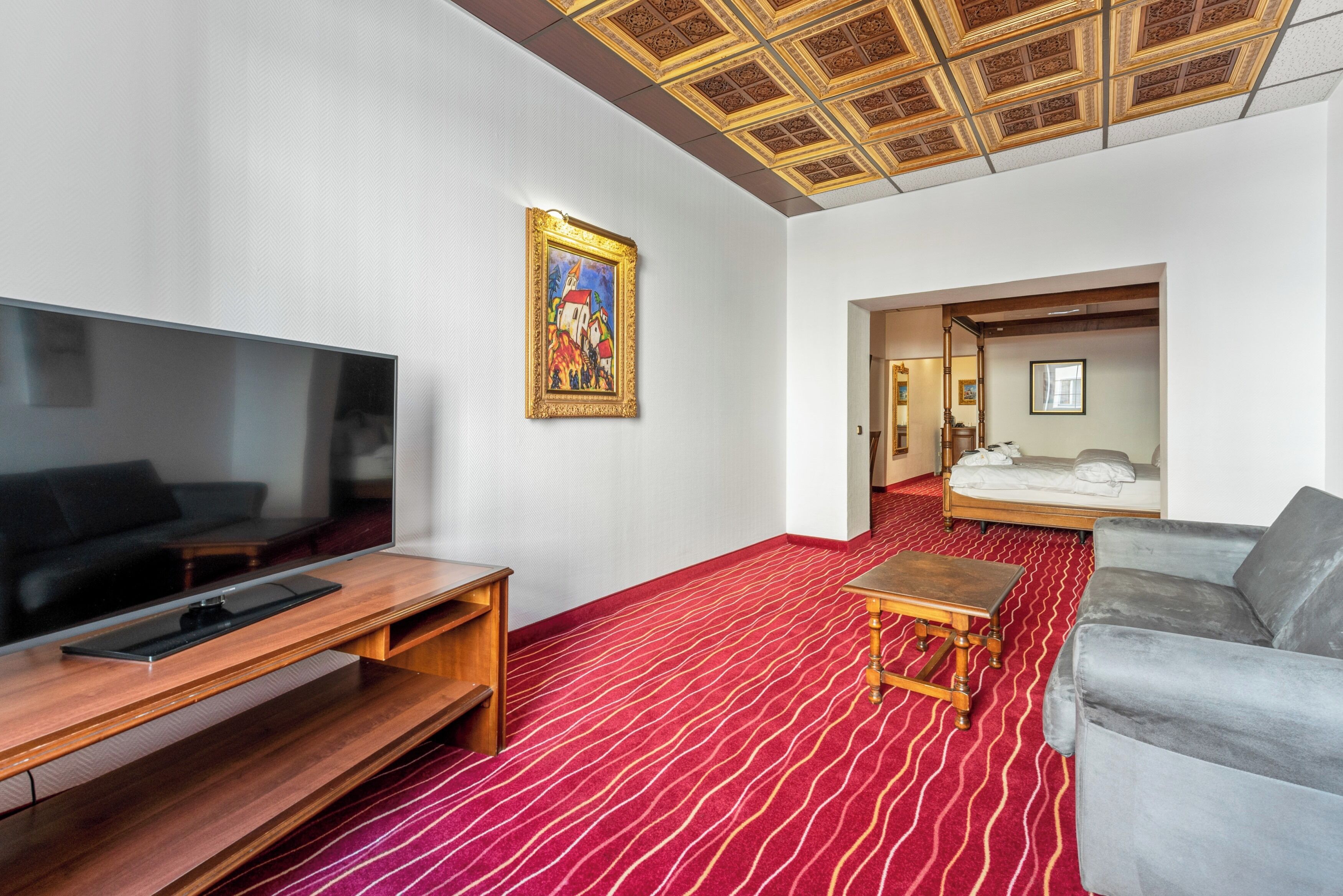 A hotel room with a large flat-screen TV, a wooden coffee table, a red patterned carpet, a gray sofa, and a ceiling with a decorative design.
