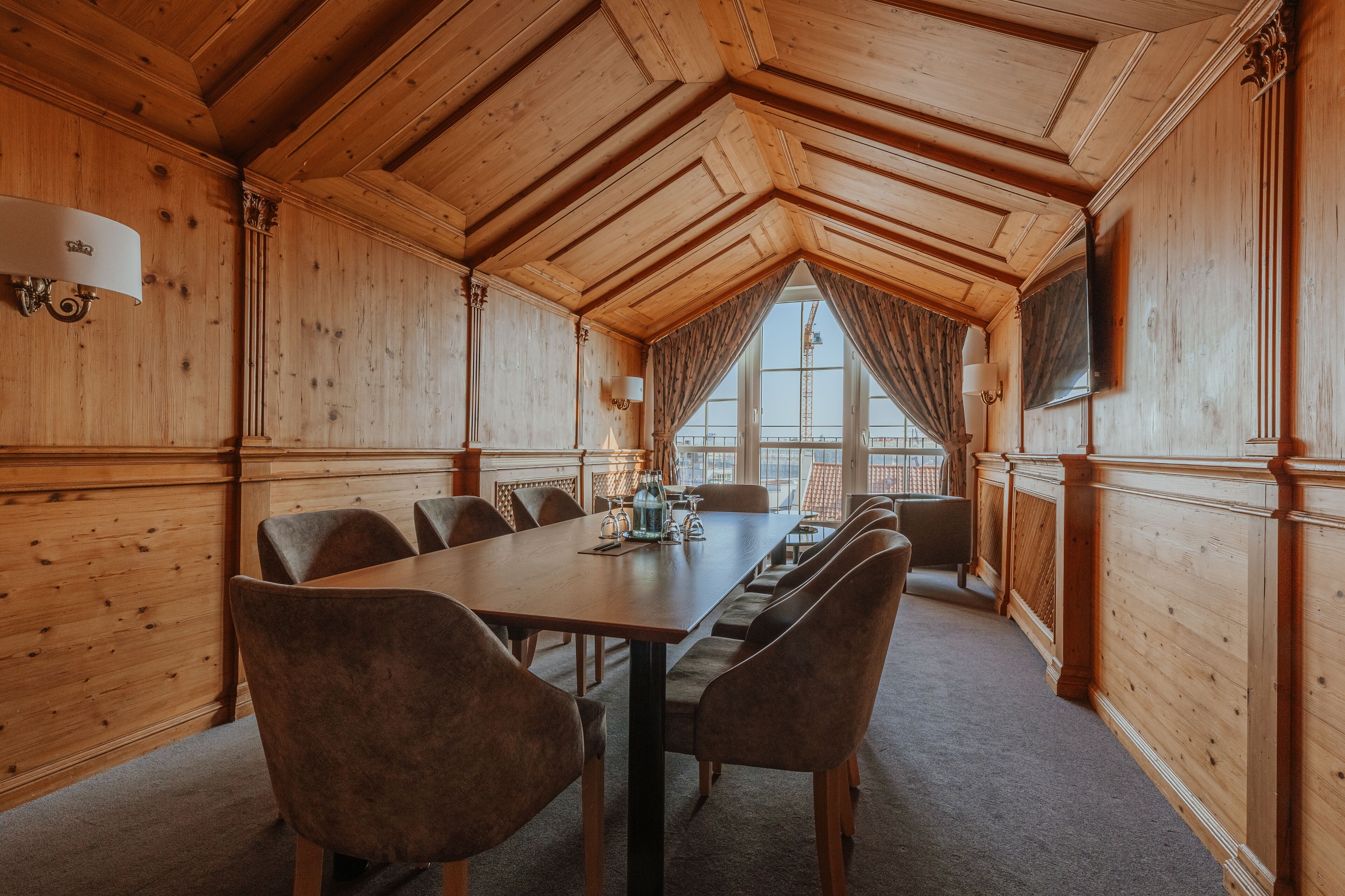 A wooden-paneled room with a long table and chairs, large windows with curtains, and a chandelier.