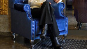 Une personne assise sur un grand fauteuil bleu orné, surmonté d’un emblème représentant une couronne, et tenant une tablette.