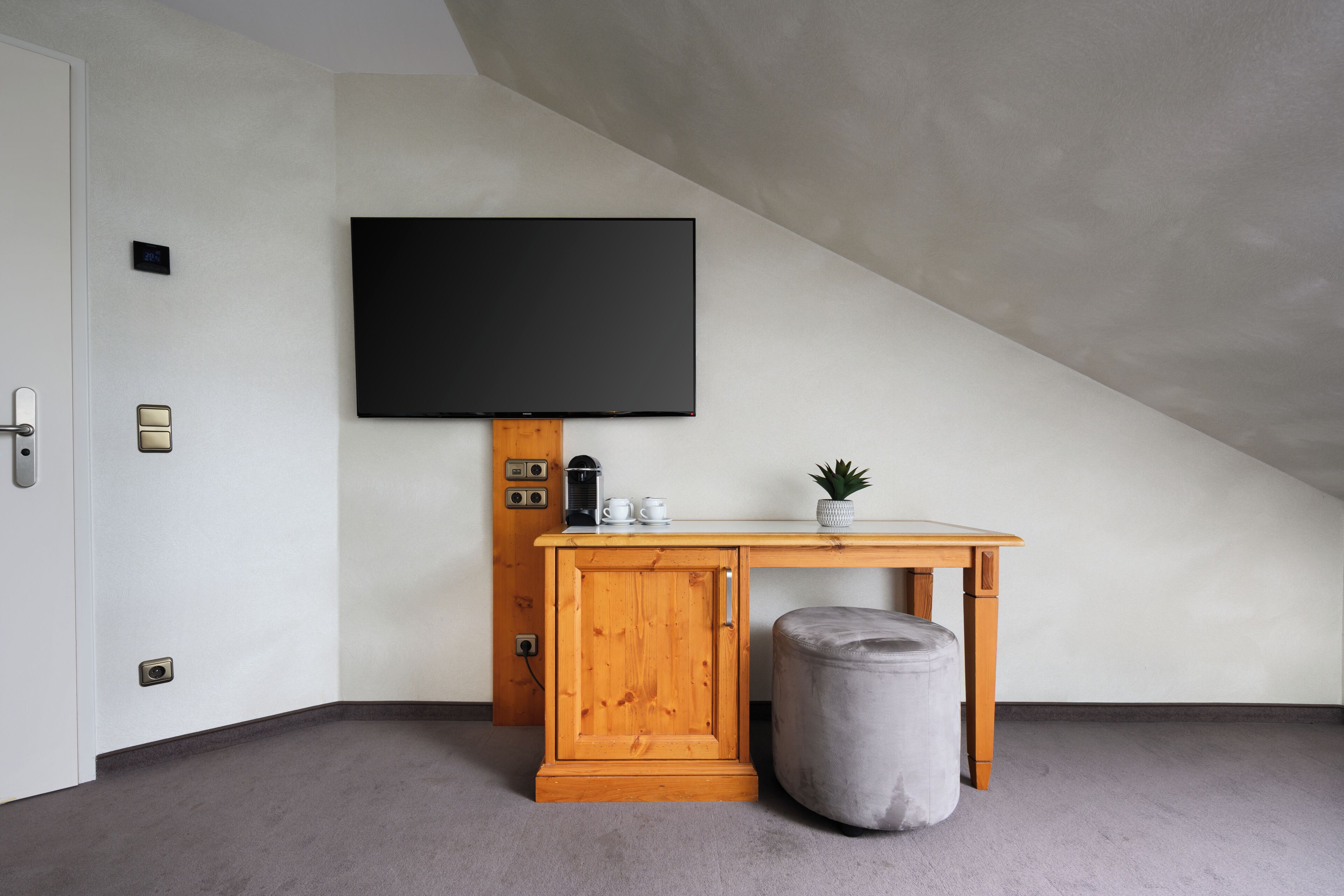 A room with a wooden cabinet, a television mounted on the wall, a small table with a plant, and a grey ottoman.