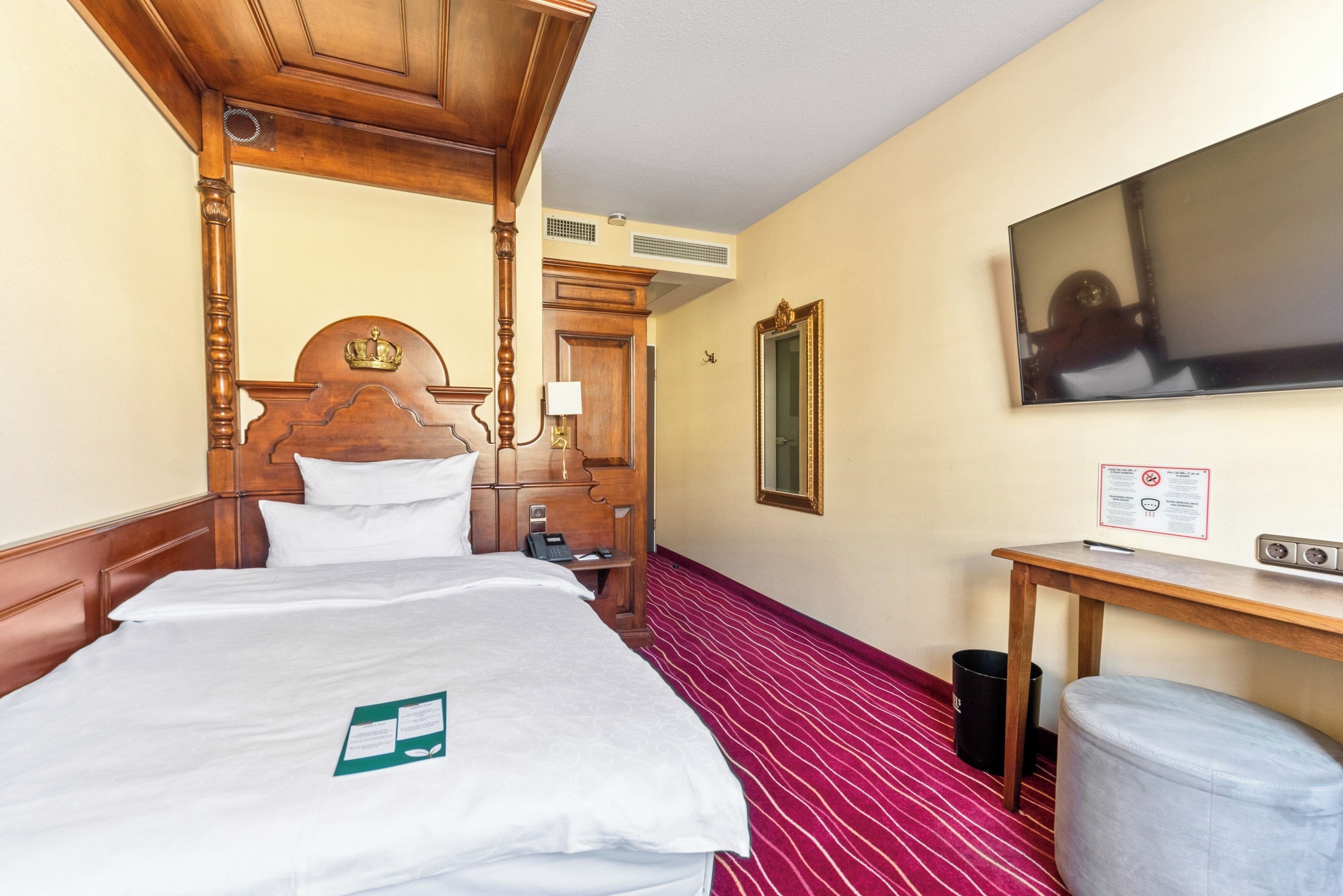 Una habitación de hotel con cama con dosel, un escritorio de madera, televisión y piso cubierto de alfombra roja.