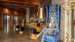Un homme est assis sur un fauteuil en velours bleu orné d’un emblème de couronne, dans un hall avec des boiseries, un canapé jaune et une lampe dorée.