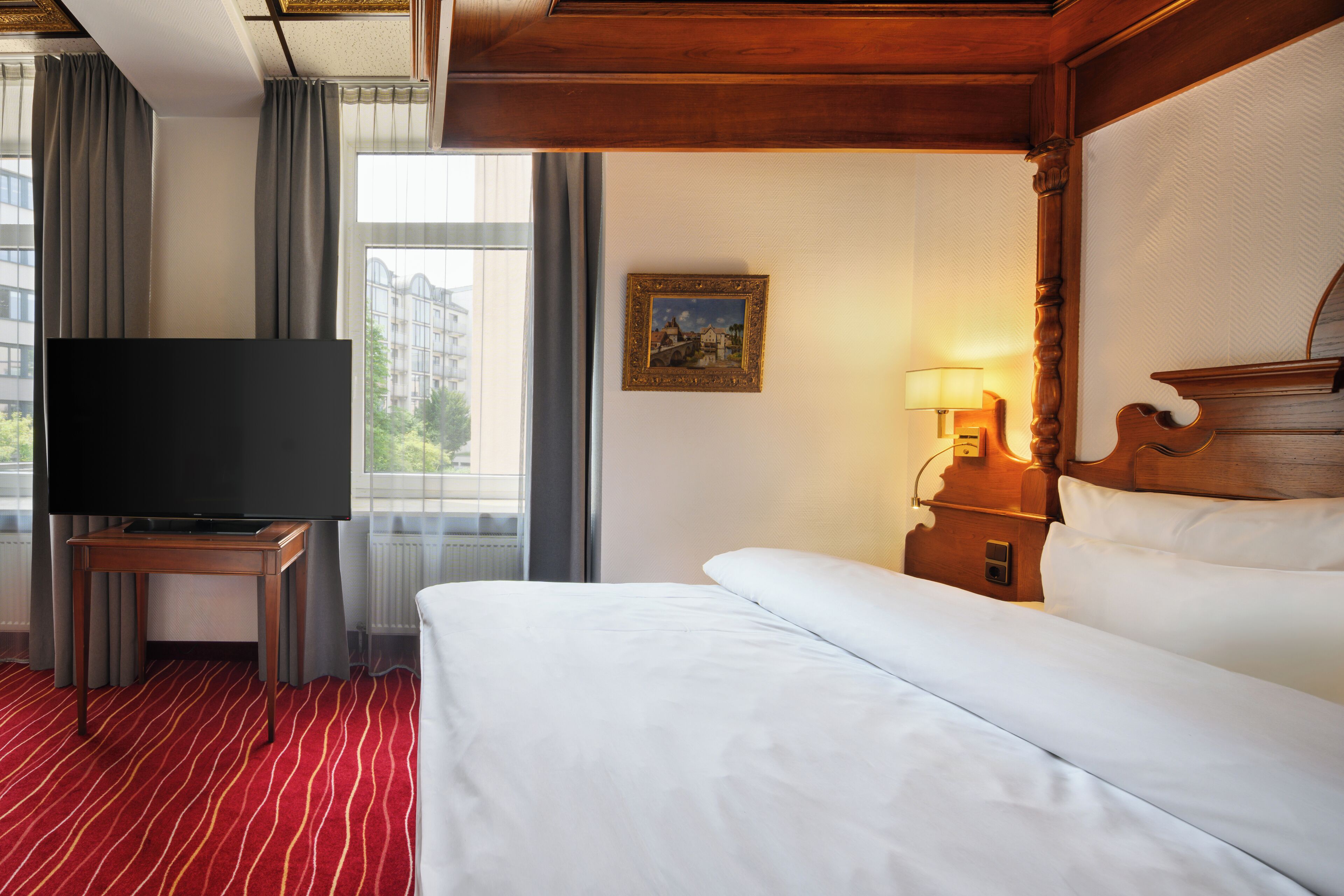 A hotel room with a large bed, a television on a wooden stand, a bedside table with a lamp, and a window with curtains.