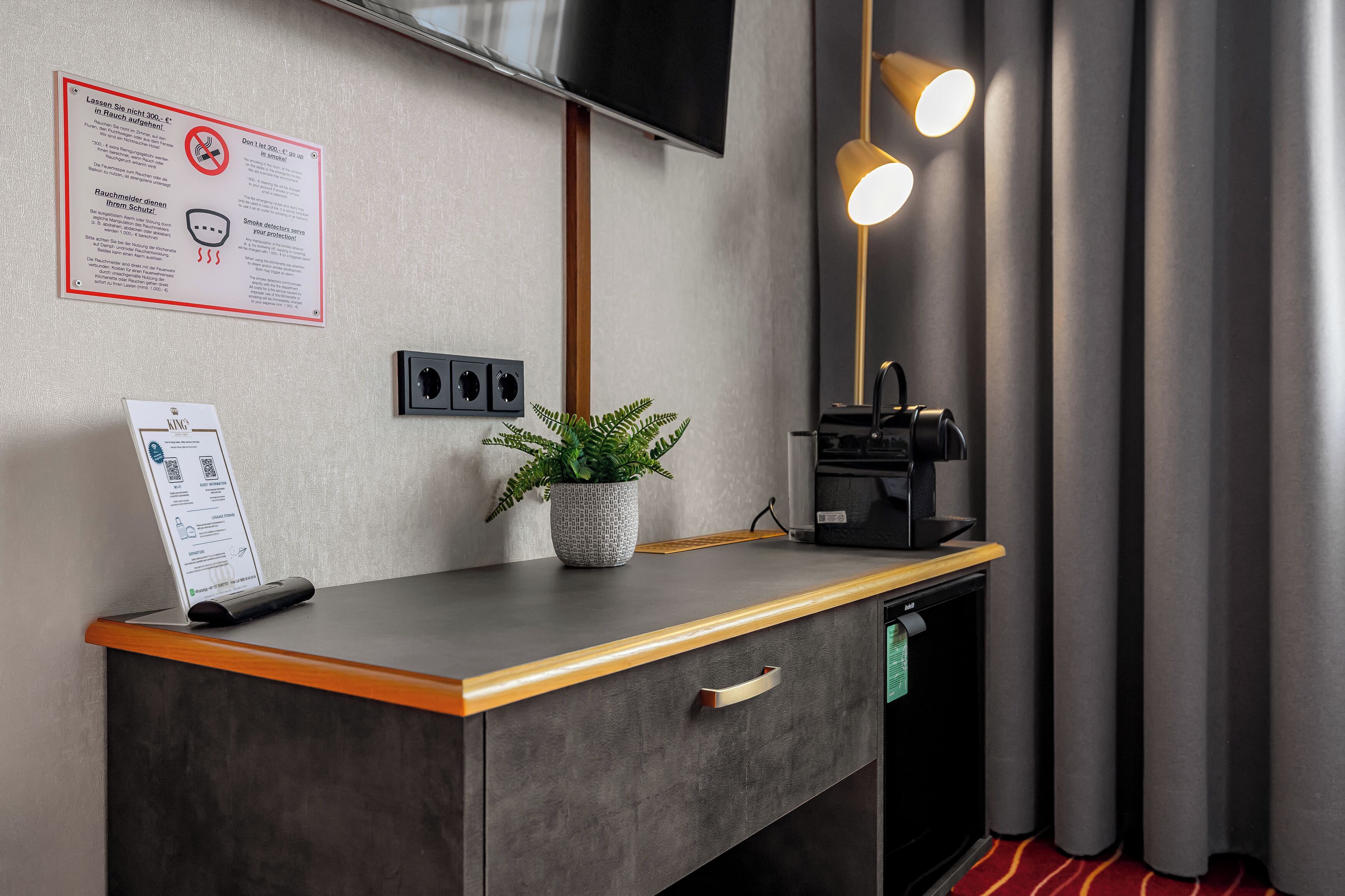 Habitación de hotel con escritorio, planta, cafetera, televisor y un cartel que indica prohibido fumar.