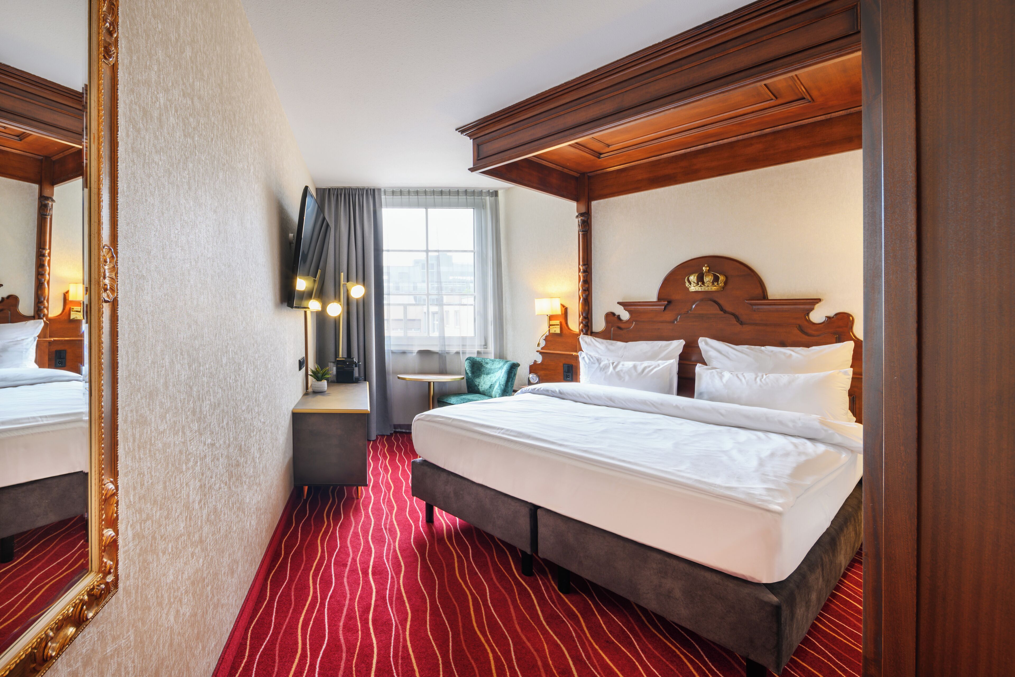 A hotel room with a large bed, a wooden headboard, a red striped carpet, a small table with a chair, and a large mirror.