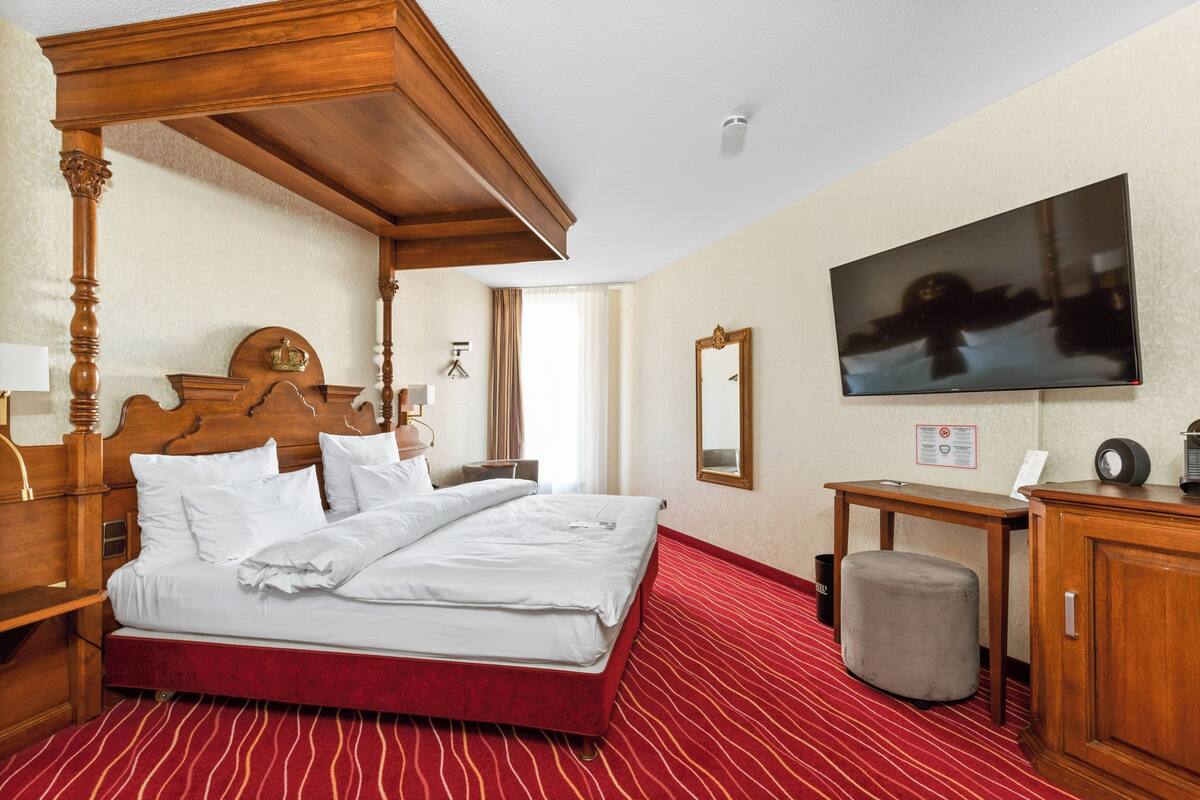 A hotel room with a four-poster bed, a flat-screen TV, a wooden nightstand, a desk, and a cabinet.