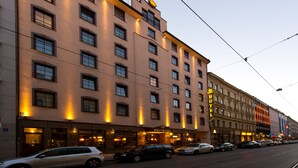 Un hôtel à plusieurs étages, reconnaissable à son grand panneau « HÔTEL », à ses nombreuses fenêtres et à la rangée de voitures garées le long de la rue.
