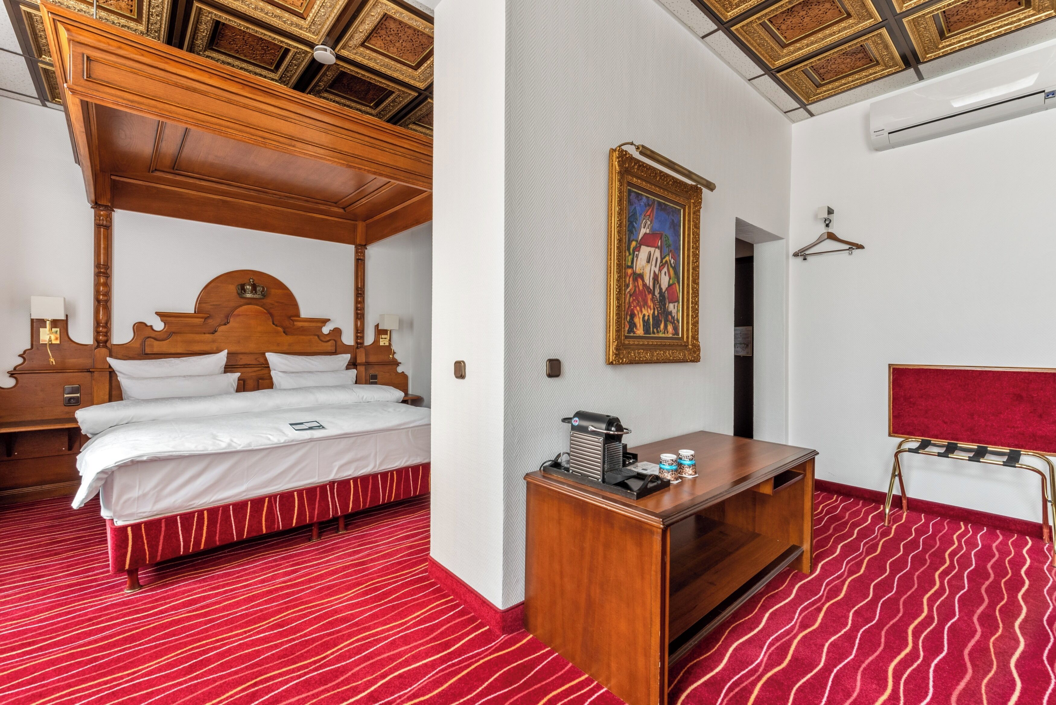 A hotel room with a four-poster bed, a wooden desk, a red patterned carpet, and a painting on the wall.