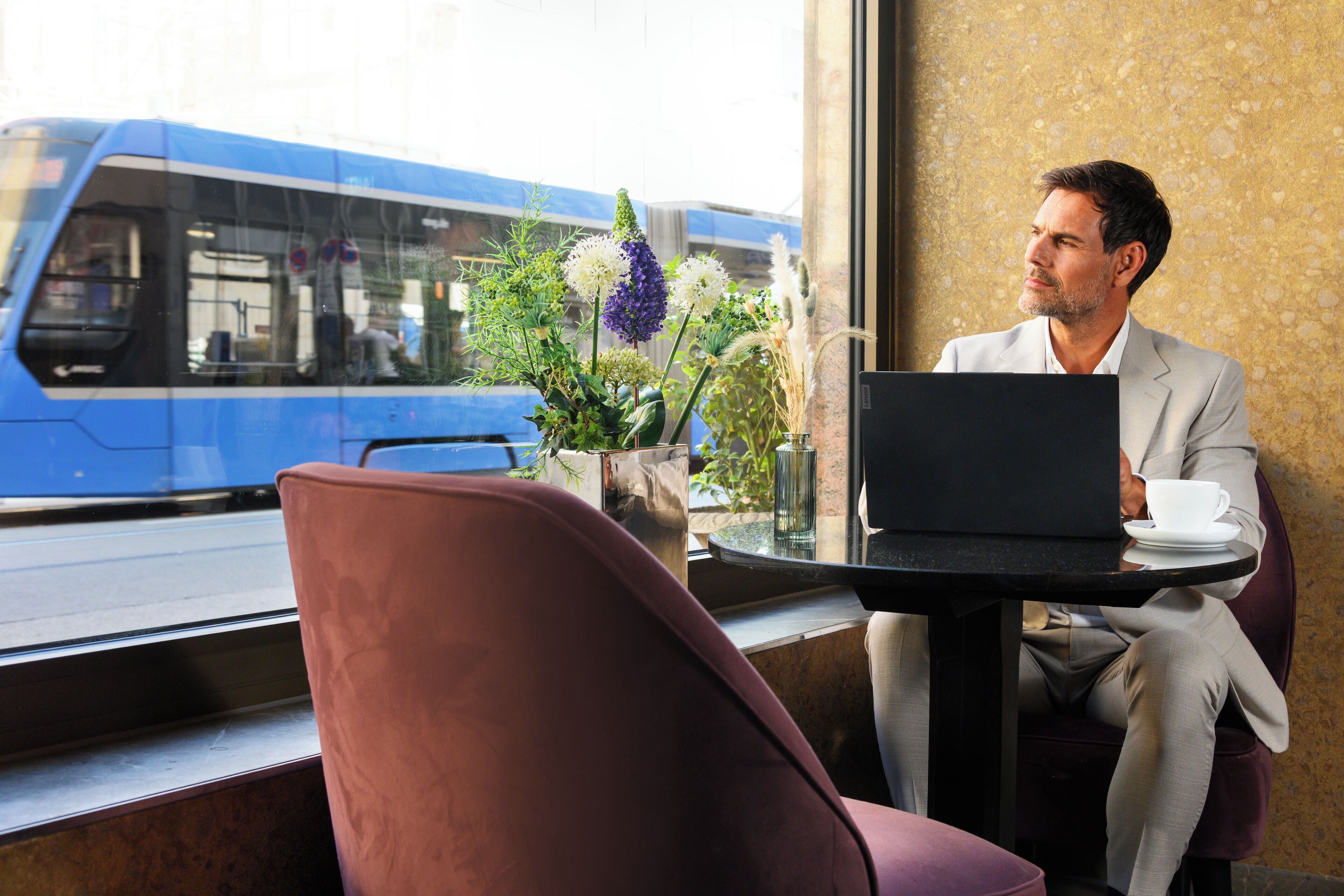 Un homme est assis à une table, devant un ordinateur portable, avec une tasse et un vase de fleurs. On aperçoit un train à travers la fenêtre.