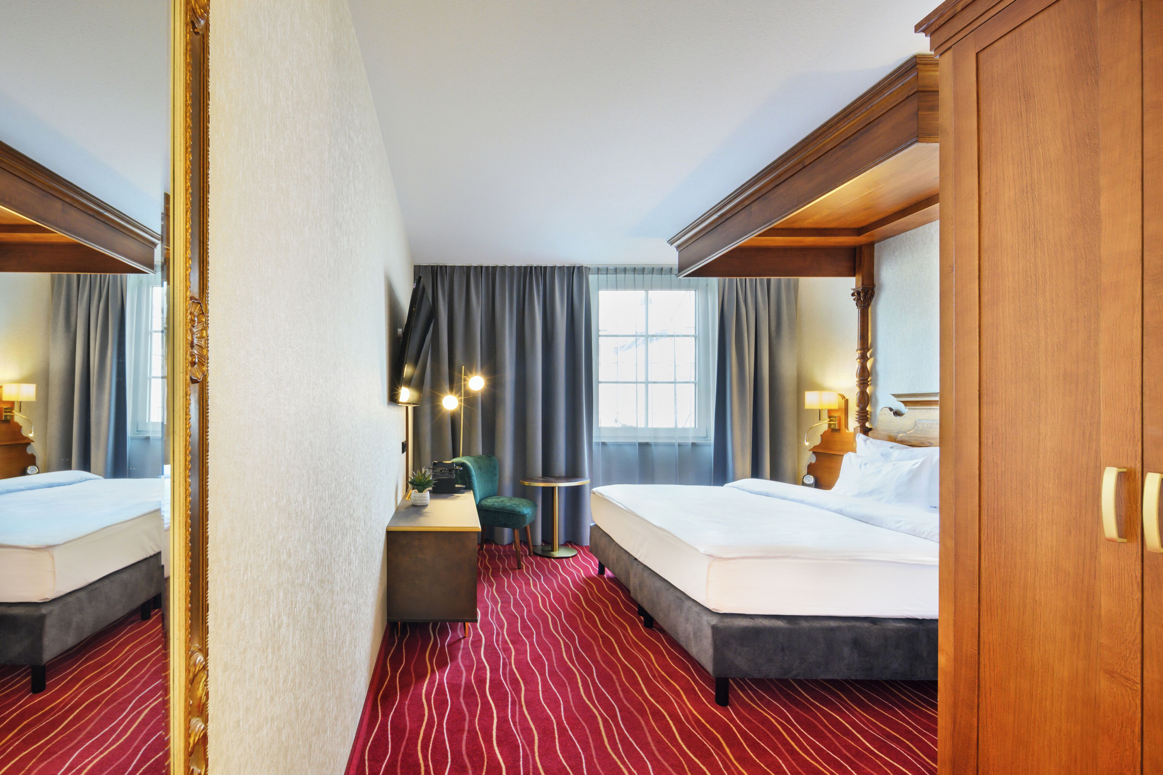 A hotel room with a large bed, a desk with a chair, a small table, and a mirror.