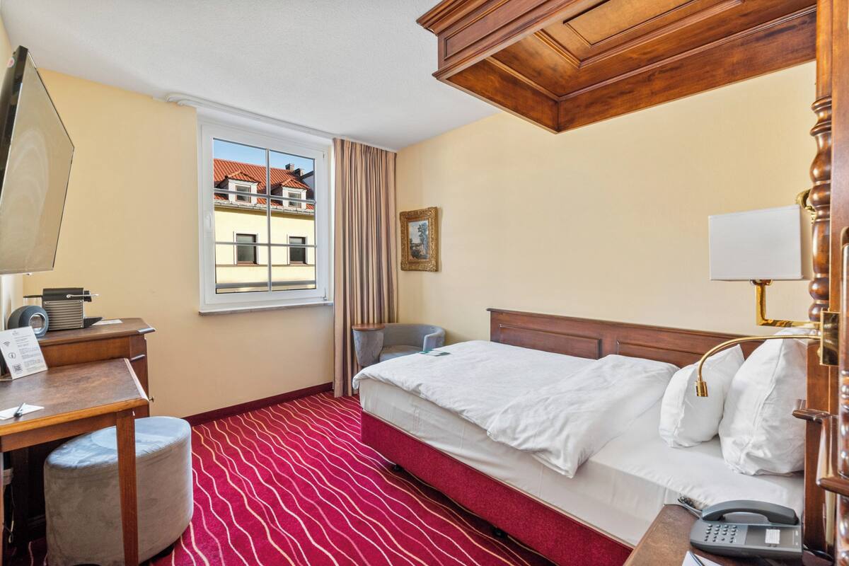 A hotel room with a bed, a desk with a telephone, a chair, a window with curtains, and a view of buildings outside.
